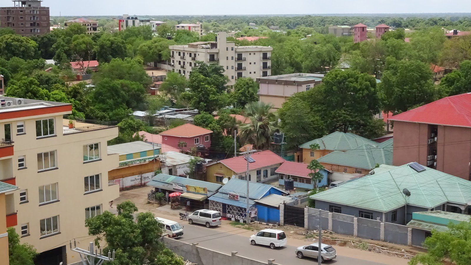Cityscape with buildings, trees, and a road with cars. Beige, red, and green buildings with lush greenery.