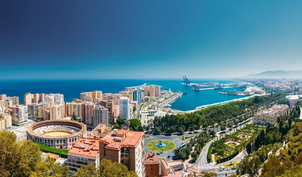 Panoramic view of Málaga, Spain, featuring city buildings, harbor, blue sea and sky, and a bullring.