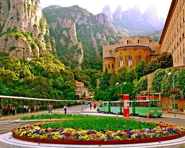 Mountainous landscape with monastery buildings, a road, flowerbed, and green buses. People are visible walking around.