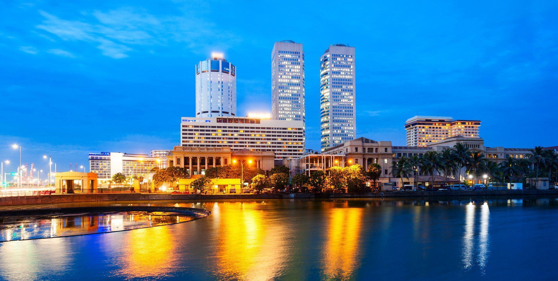 Nighttime cityscape view of Colombo, Sri Lanka, with lit-up high-rise buildings reflecting in the calm water.