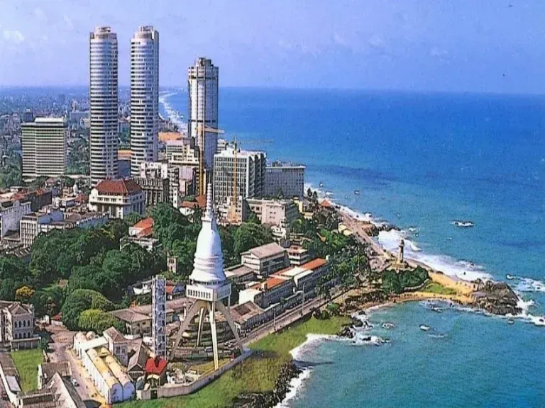 Cityscape of Colombo, Sri Lanka, with modern skyscrapers and a coastal view, including a prominent white clock tower and the blue ocean.