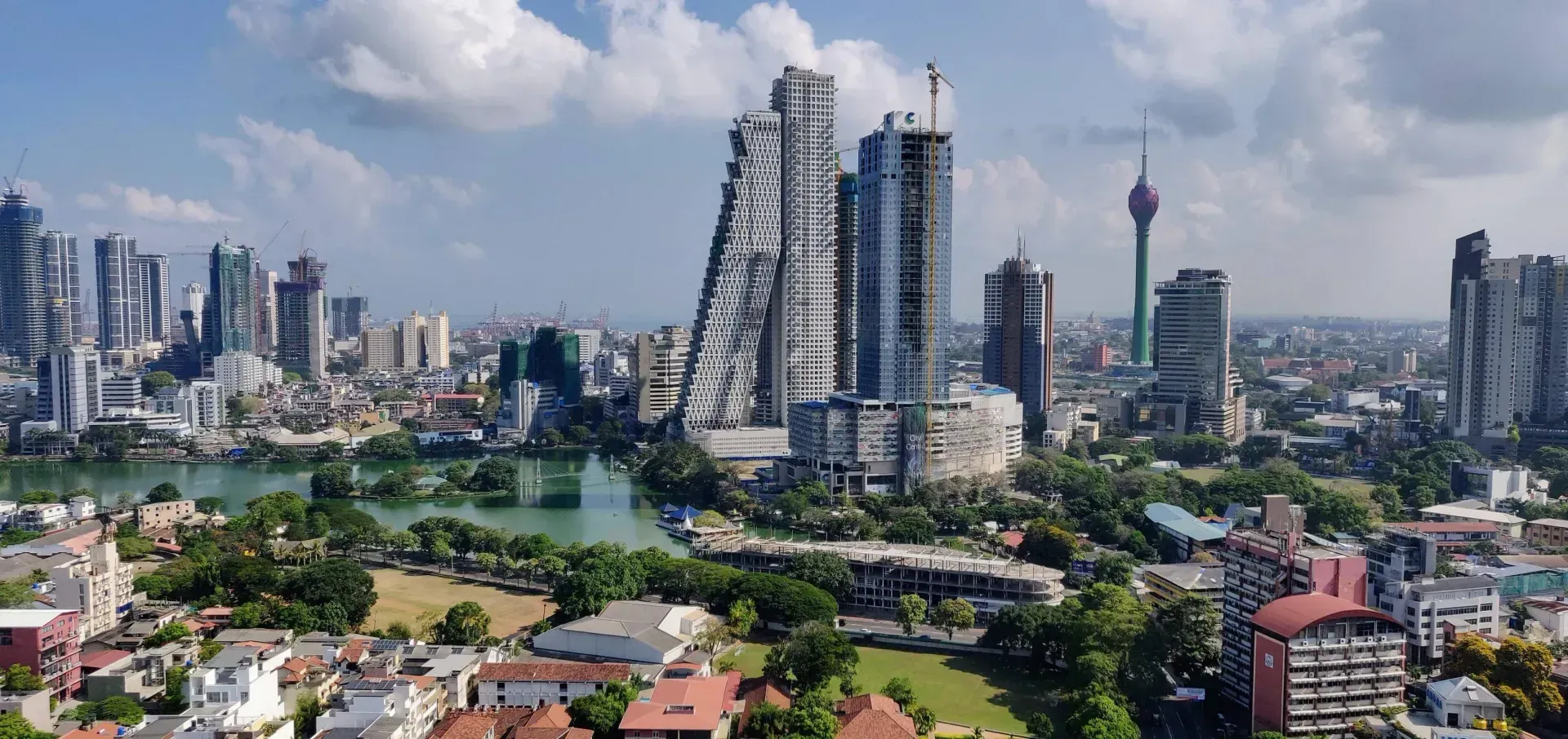 City skyline with tall buildings, a lake, and a tower with a distinctive shape, possibly Colombo, Sri Lanka.