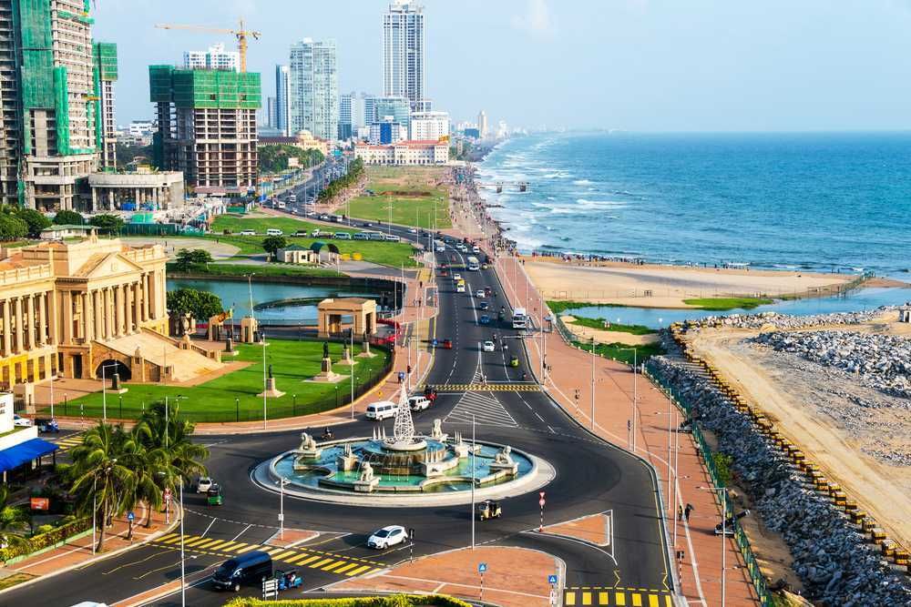 Coastal city view in Sri Lanka: A road curves along the ocean, passing a fountain, with buildings, beach, and blue sea.