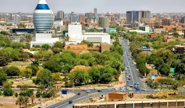 A cityscape in Khartoum, Sudan, with a tall tower, wide road, trees, and buildings under a bright sky.