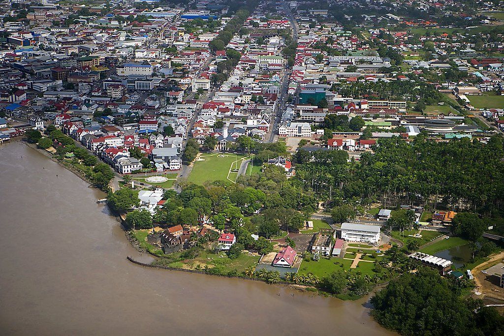 Aerial view of Georgetown, Guyana, with a brown river bordering the city, and rows of buildings and green spaces extending inland.