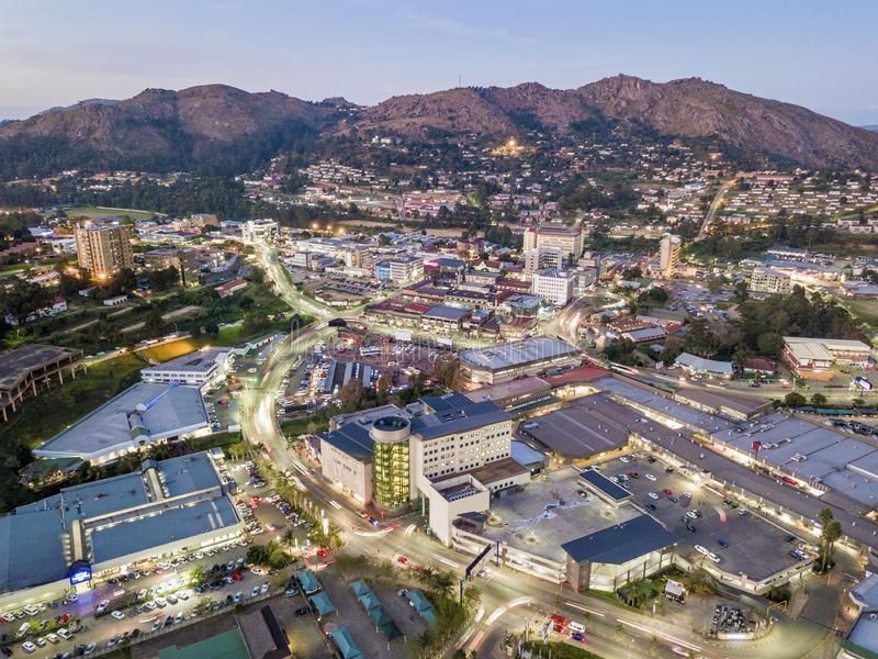 Aerial view of a city at dusk with buildings, roads, and mountain backdrop. Lights are on in buildings and along streets.
