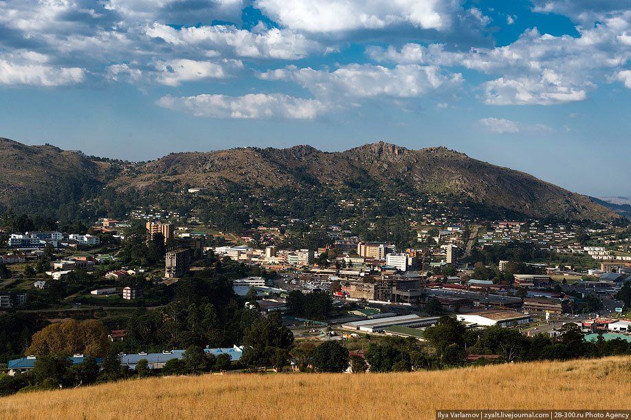 Town nestled at the base of a tree-covered mountain, under a blue sky with white clouds.