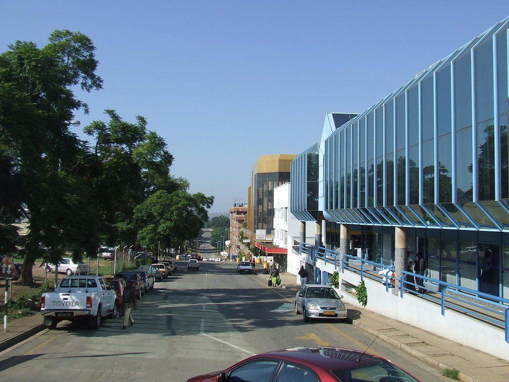 Street scene in a city with cars, trees, and modern buildings. Blue glass facade on the right, clear sky overhead.