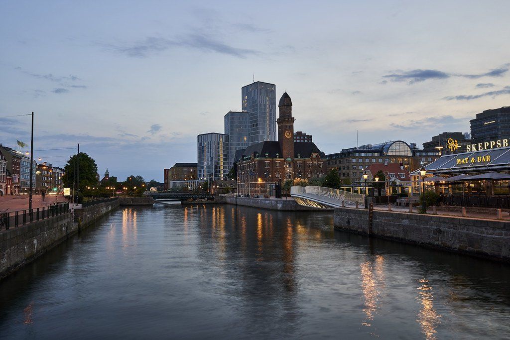 A twilight cityscape with a canal. Tall buildings reflect in the water, along with lights from the shore.