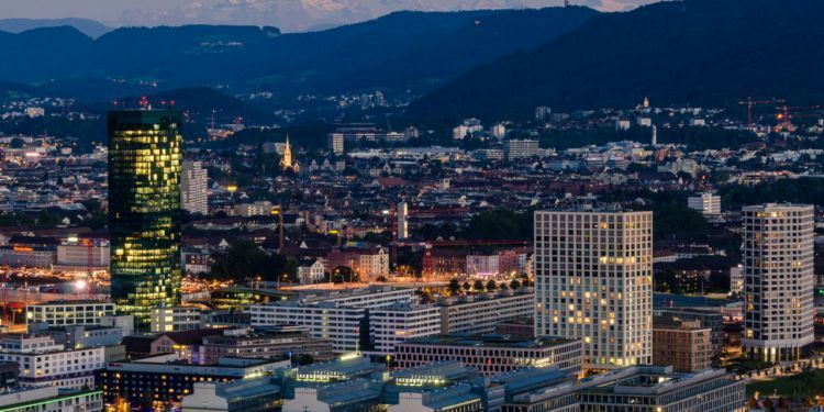 Cityscape at dusk with illuminated skyscrapers and mountains in the background. Buildings and city lights glow against a dark blue sky.