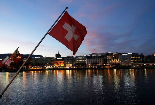 Swiss flag waving over a lake at dusk, with city lights reflecting in the water.