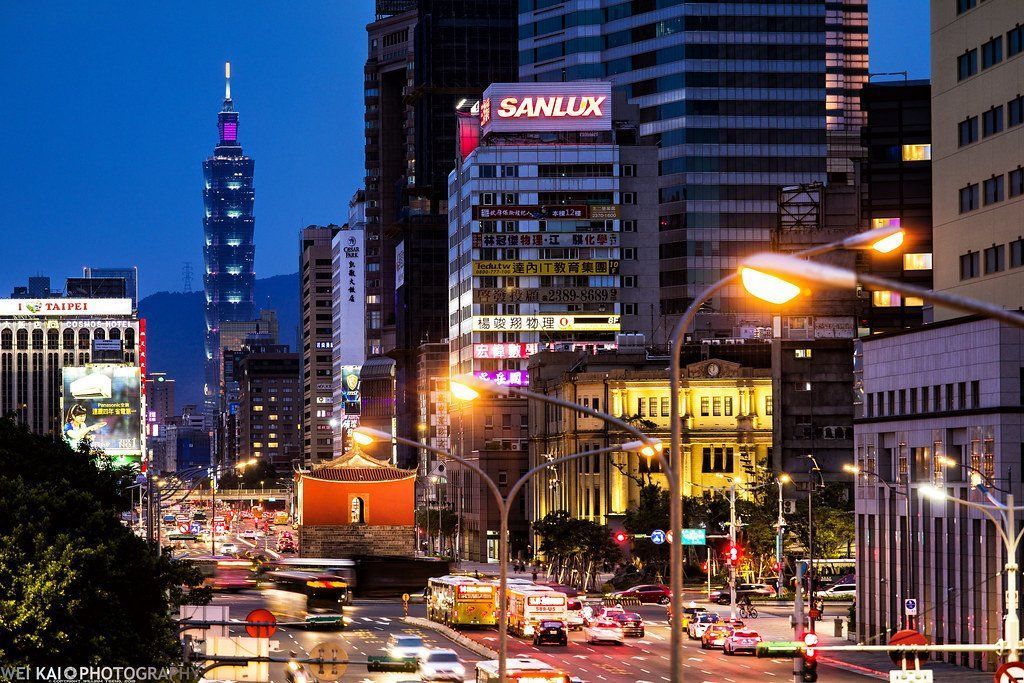 Nighttime cityscape of Taipei, Taiwan, with illuminated buildings and traffic on a busy road; Taipei 101 visible in the background.