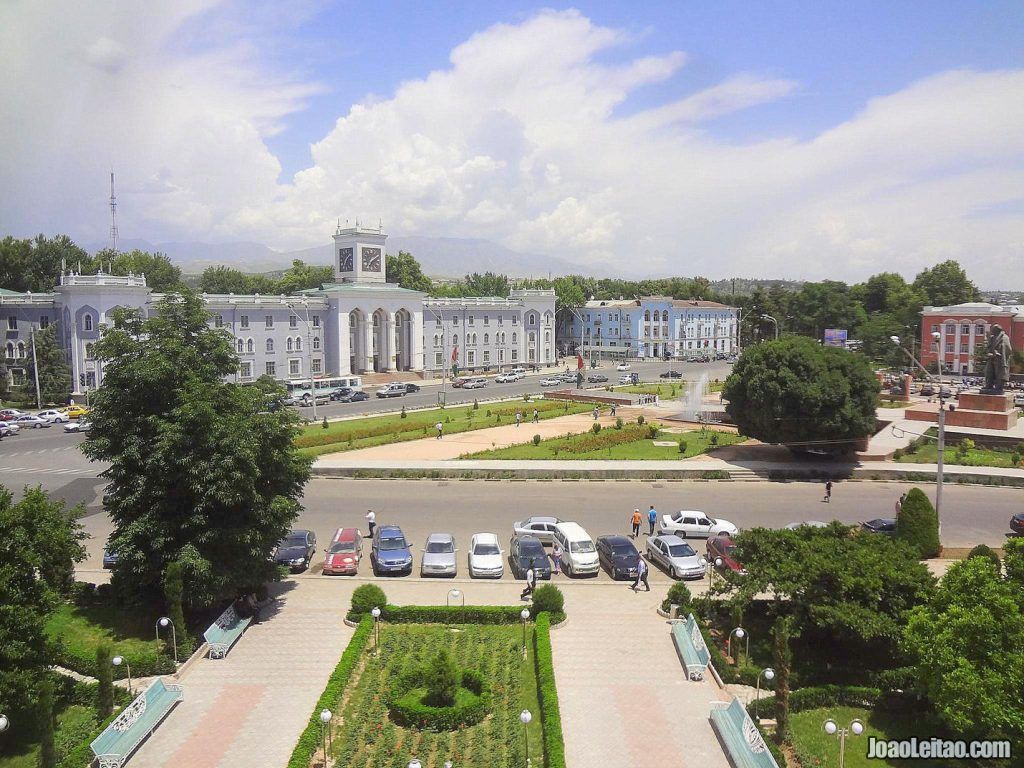 View of a city square with government buildings, trees, and parked cars under a blue sky with white clouds.