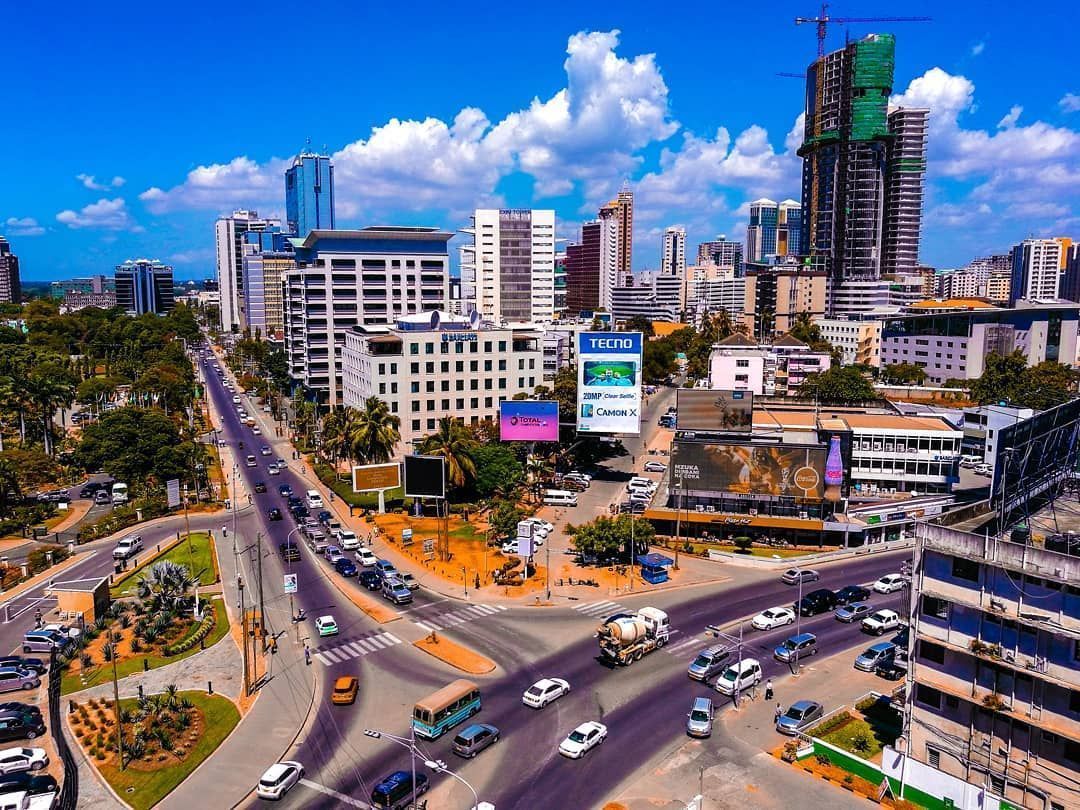 Cityscape with a wide road intersection, featuring tall buildings, traffic, and construction; blue sky.