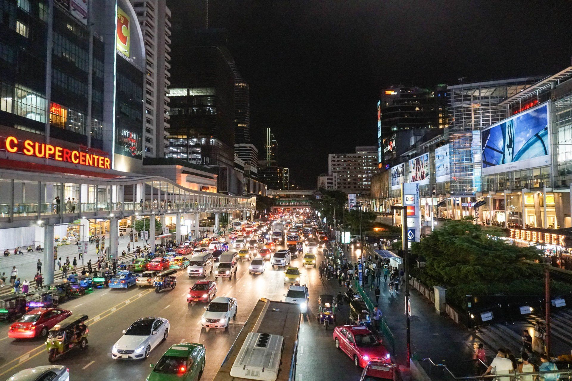 Nighttime cityscape in Bangkok, Thailand. Busy street filled with cars and pedestrians, lined by brightly lit buildings.