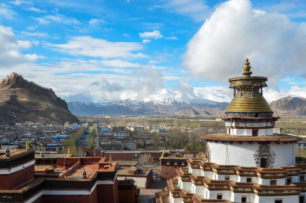 View of a white and gold stupa with a city and snow-capped mountains in the background under a blue sky.