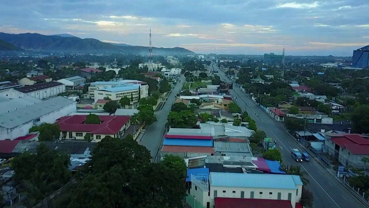 Aerial view of a town with a central road, buildings, and mountains in the background under a cloudy sky.