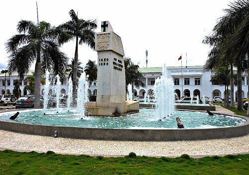 Fountain with monument in the center, surrounded by jets of water. Palm trees and a white building are in the background.
