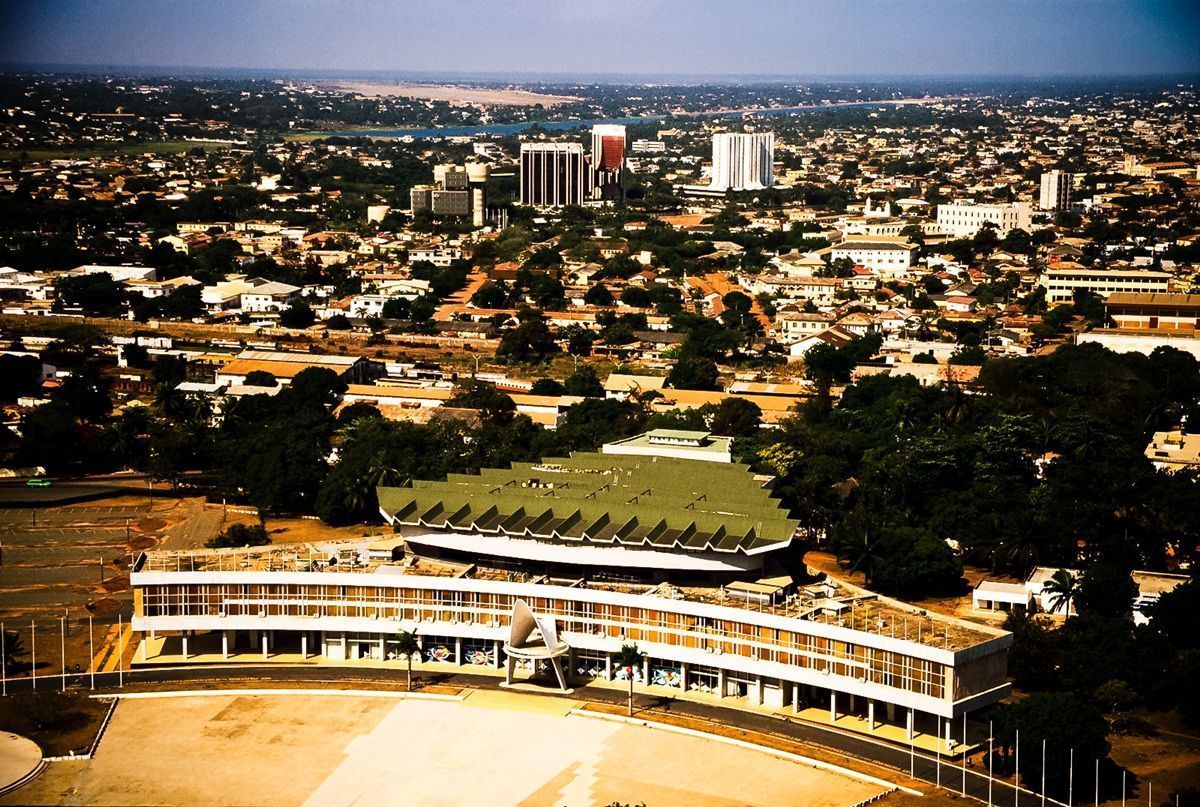 Aerial view of a city, possibly African, with a unique, tiered building in the foreground. The sky is overcast.