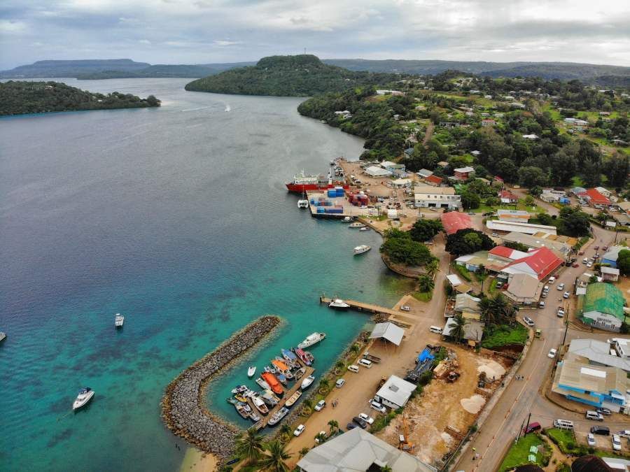 Aerial view of a coastal town with colorful buildings, boats in a harbor, and a lush green hillside in the background.