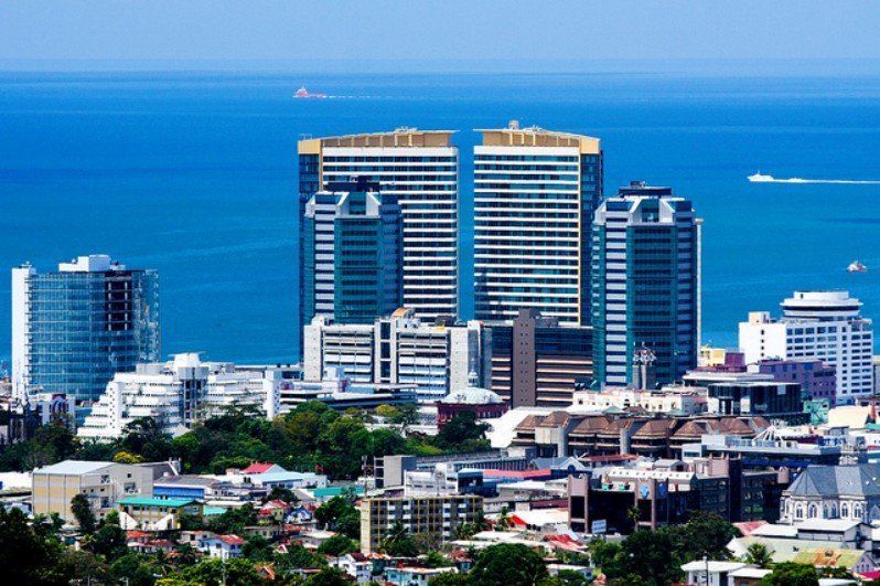 City skyline with modern high-rise buildings against a blue ocean backdrop. The sky is clear, and some boats are visible in the distance.