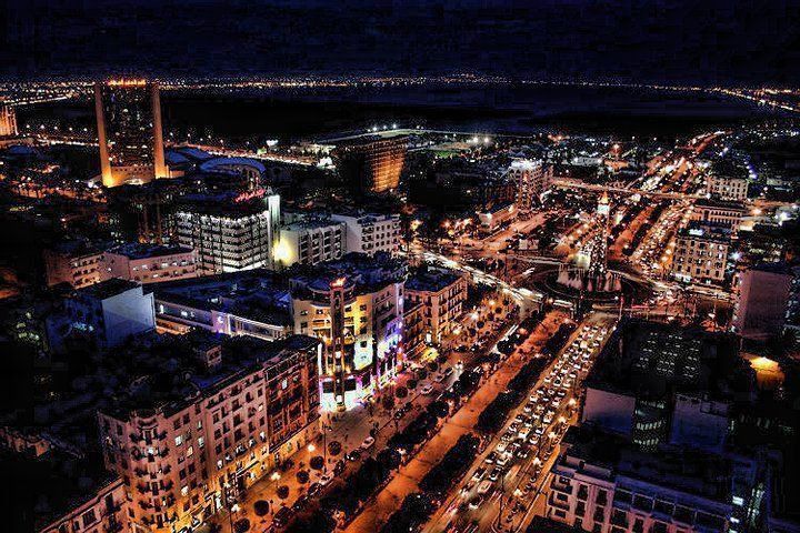 Nighttime aerial view of a brightly lit city with roads, buildings, and tall structures against a dark sky.