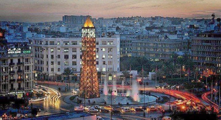 Lit-up clock tower with a fountain in a city square at dusk; cars streak around the traffic circle.