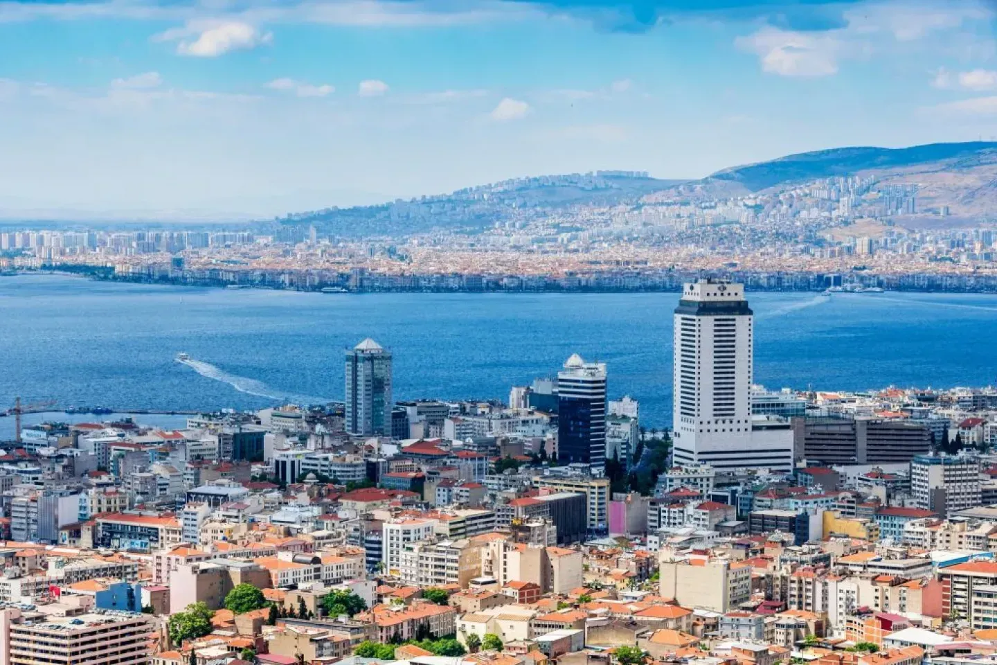 Cityscape of İzmir, Turkey, featuring buildings along the coast and a wide blue sea with mountains in the background.