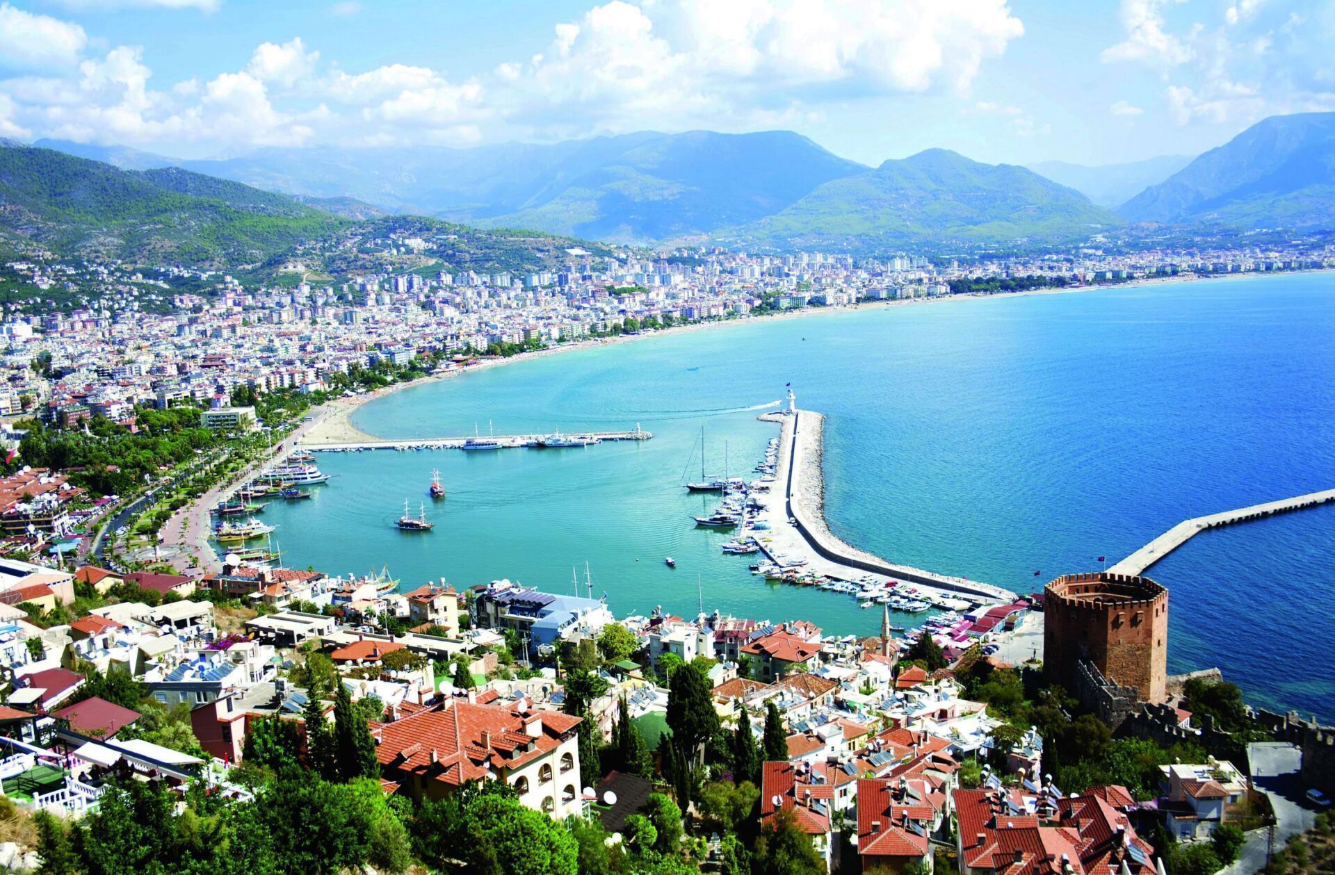 Coastal city with turquoise water, a long pier, and mountains in the background under a bright sky. Buildings with red roofs and a stone tower are in the foreground.