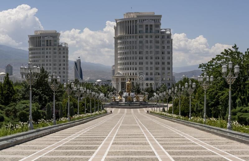 A wide, symmetrical walkway leads toward two large, white buildings in a city. Lush greenery and ornate lampposts line the path.