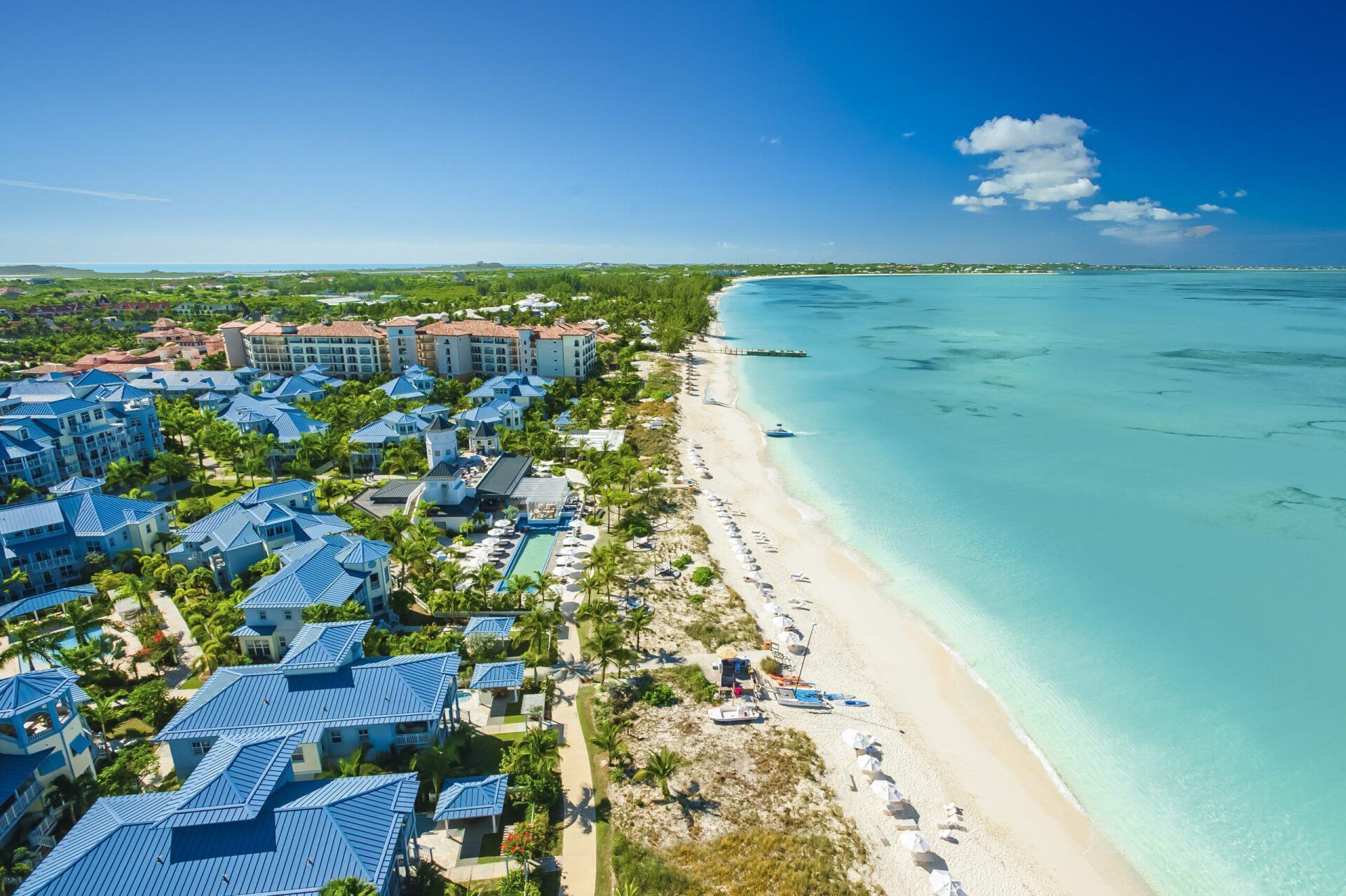 Aerial view of a luxurious beach resort with blue rooftops, white sand, and turquoise ocean under a bright blue sky.
