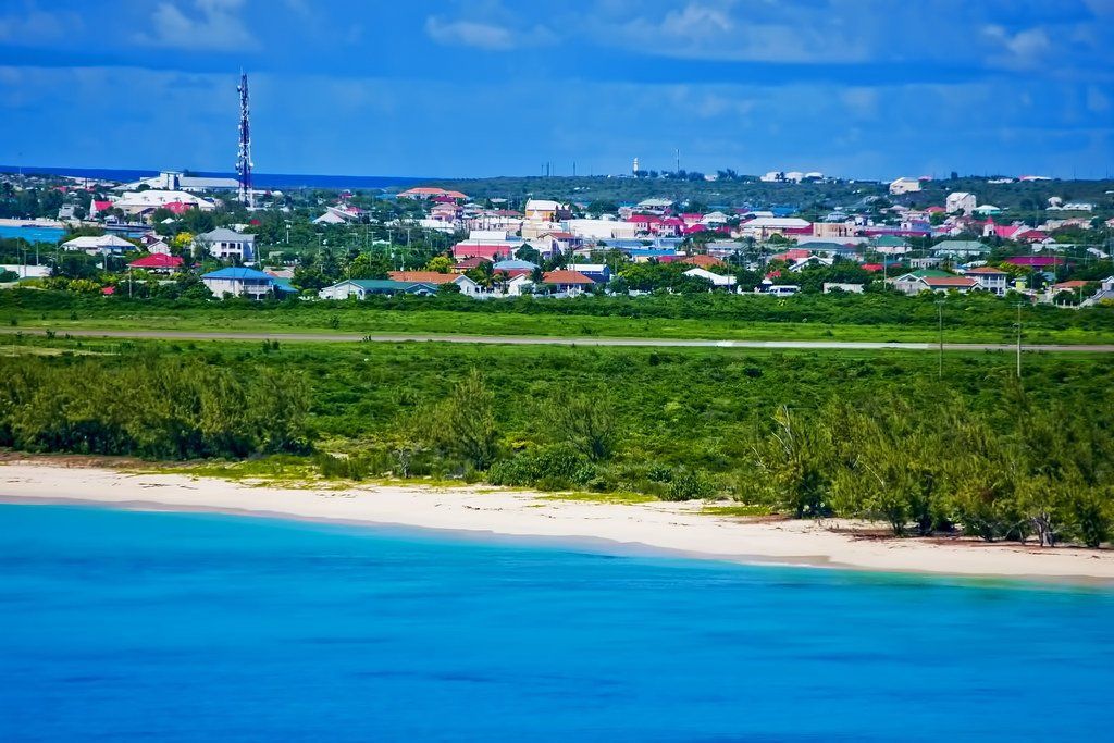 A coastal cityscape with turquoise water, a sandy beach, green trees, and buildings under a blue sky.