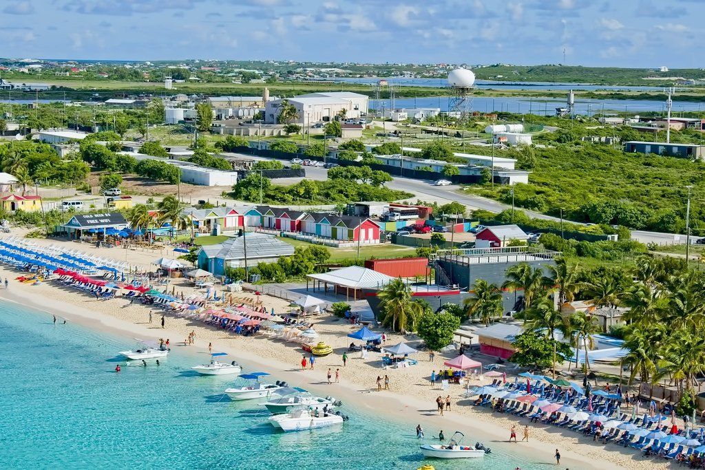 Beach with people, boats, and colorful buildings. Lush greenery surrounds the shoreline with a town in the background.