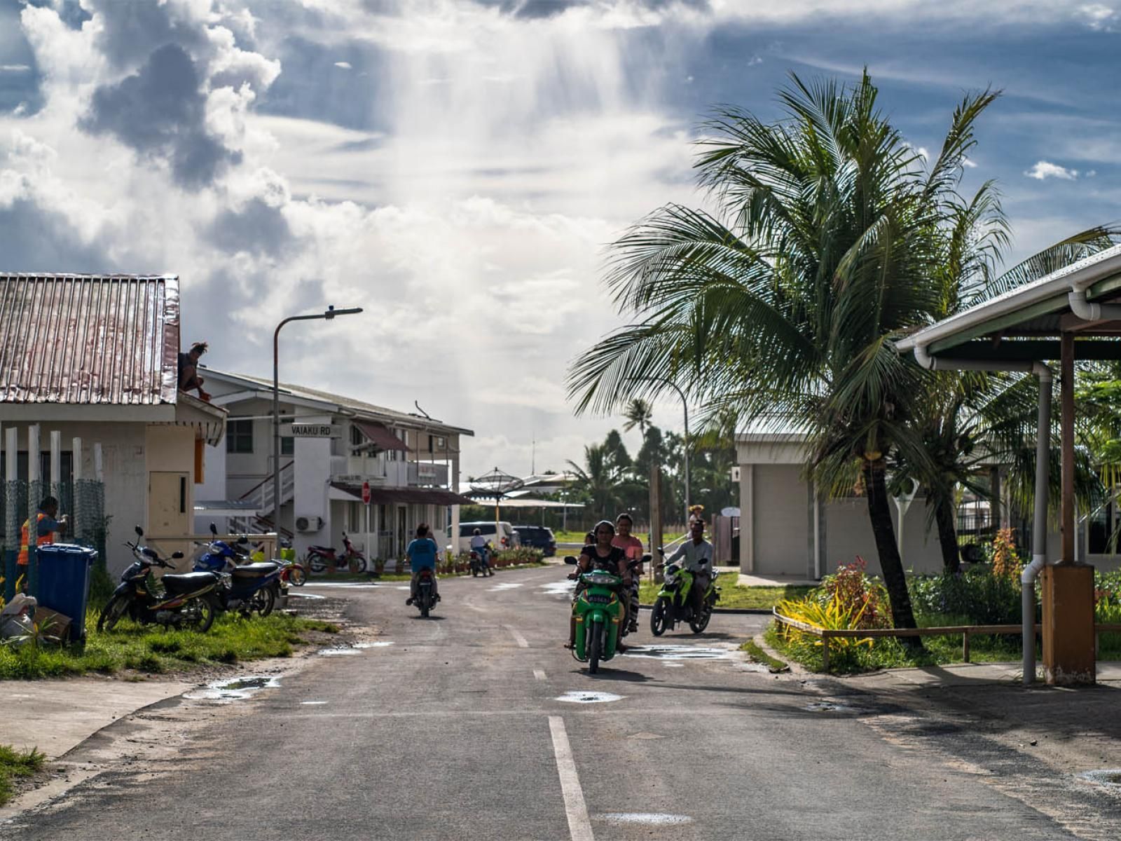 Street scene in a tropical area; people on scooters travel down a road lined with buildings and palm trees. The sky is overcast.