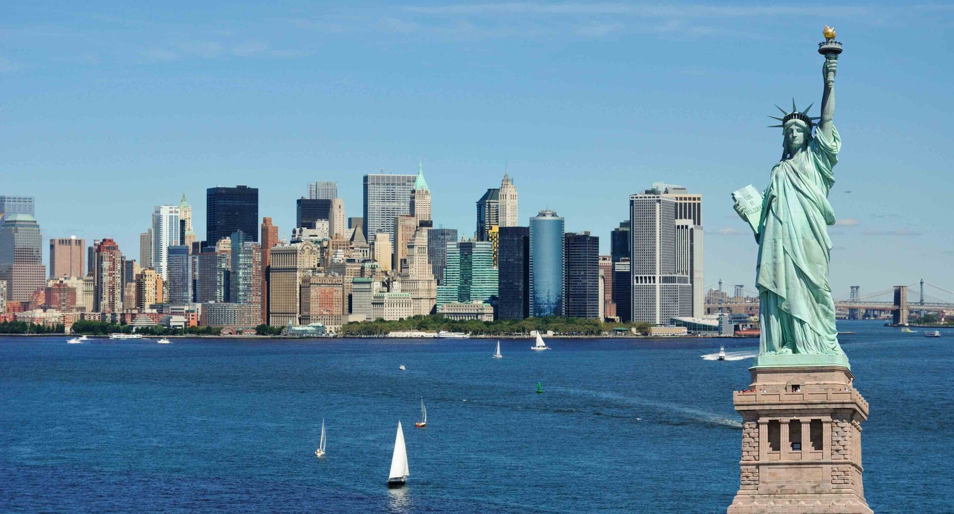 Statue of Liberty with NYC skyline in the background on a sunny day; boats sail on the water.