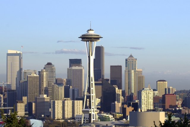 Seattle skyline with the Space Needle prominent against a blue sky; various skyscrapers fill the background.