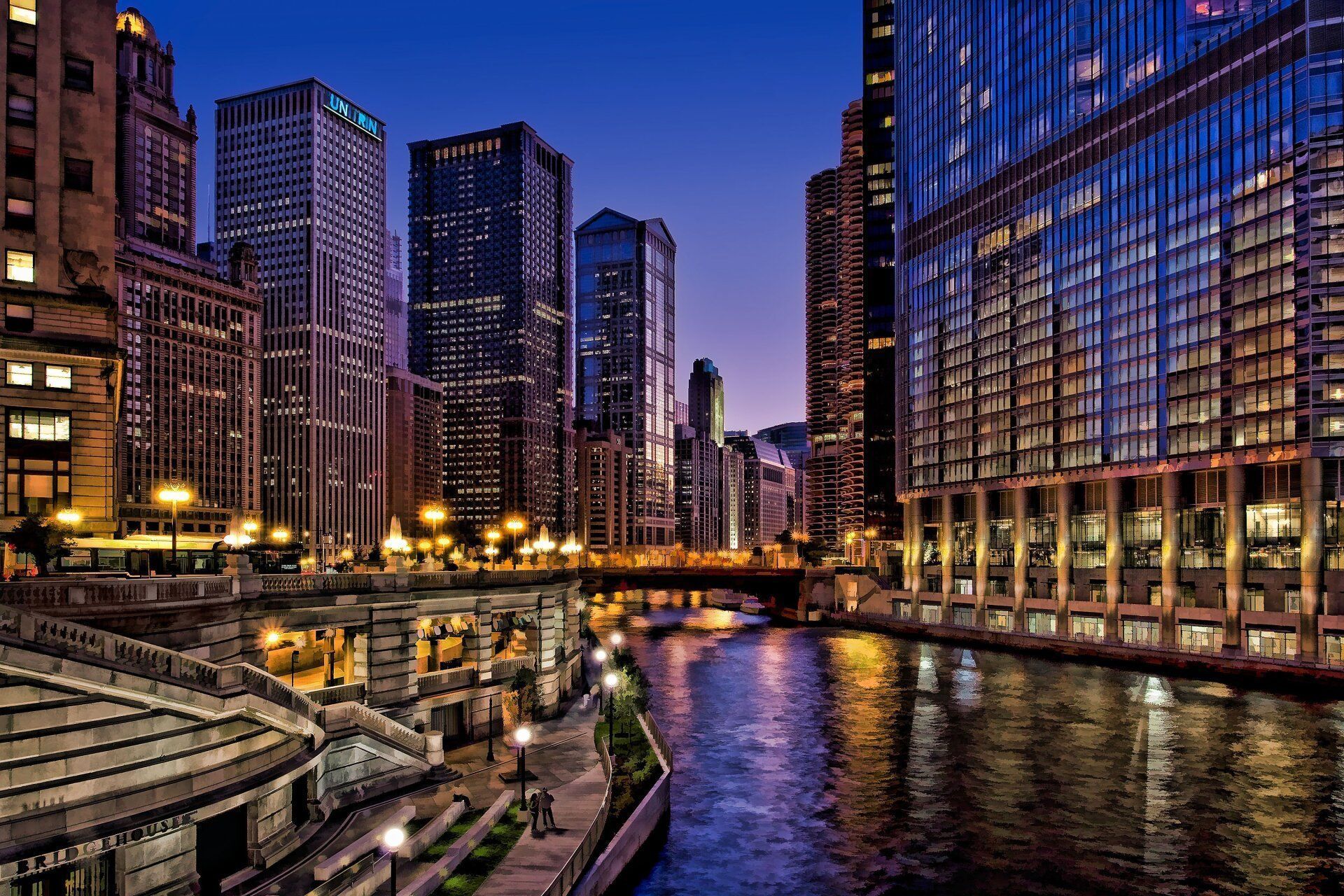 Chicago River at dusk, lined with illuminated skyscrapers. Reflective blue water and city lights create a vibrant nighttime cityscape.