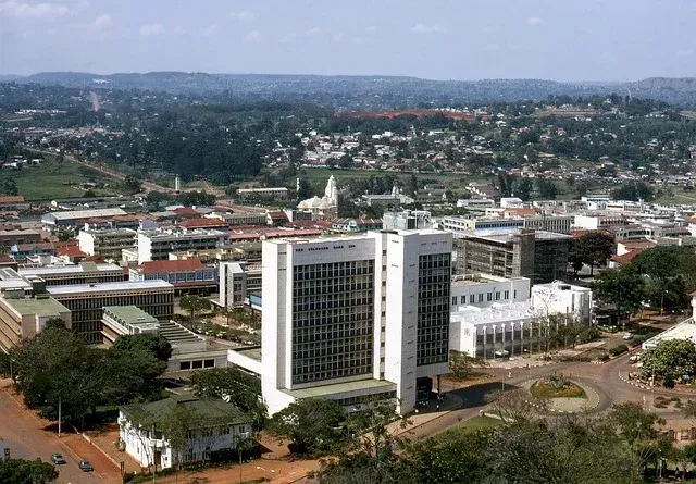 Aerial view of a cityscape with a large white building in the foreground, green trees, and buildings of various heights scattered throughout.
