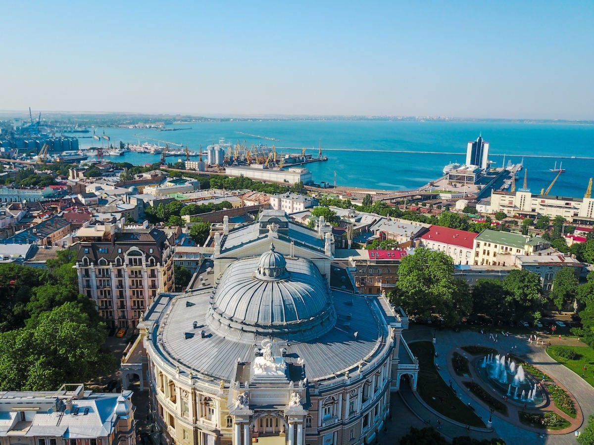 Aerial view of the Odessa Opera and Ballet Theater with a cityscape and the Black Sea in the background under a clear blue sky.