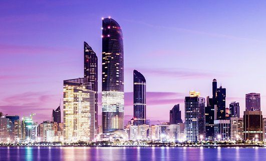 Nighttime cityscape of Abu Dhabi with illuminated skyscrapers reflecting in the water, under a purple sky.
