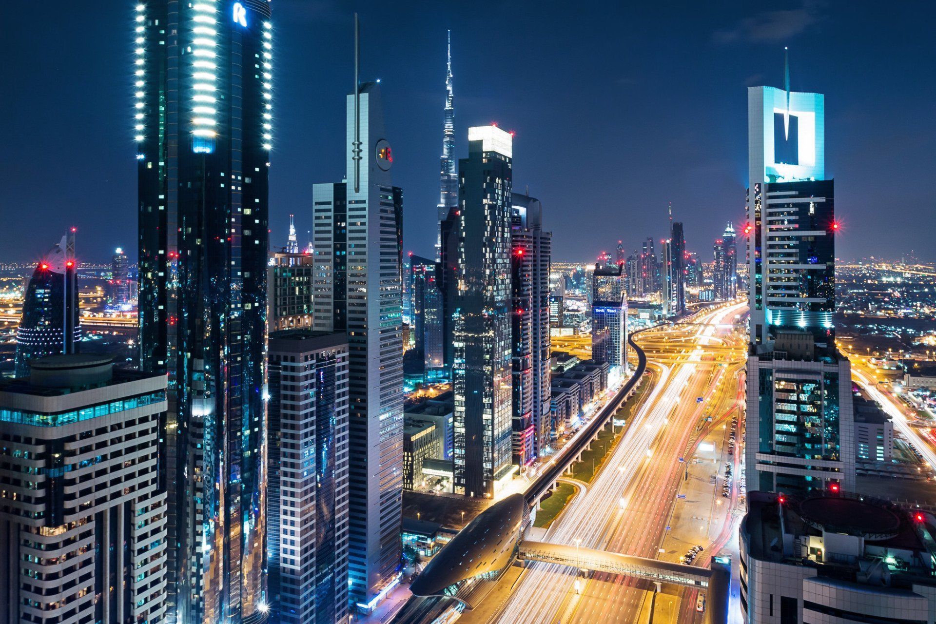 Night view of Dubai's illuminated skyscrapers and roadways. Long exposure shows streaks of car headlights on the highway.