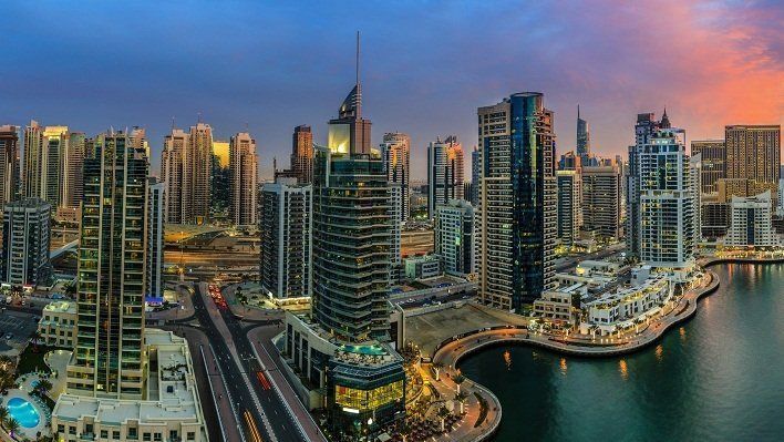 Dubai skyline at dusk with skyscrapers, a waterway, and a colorful sunset in the background.