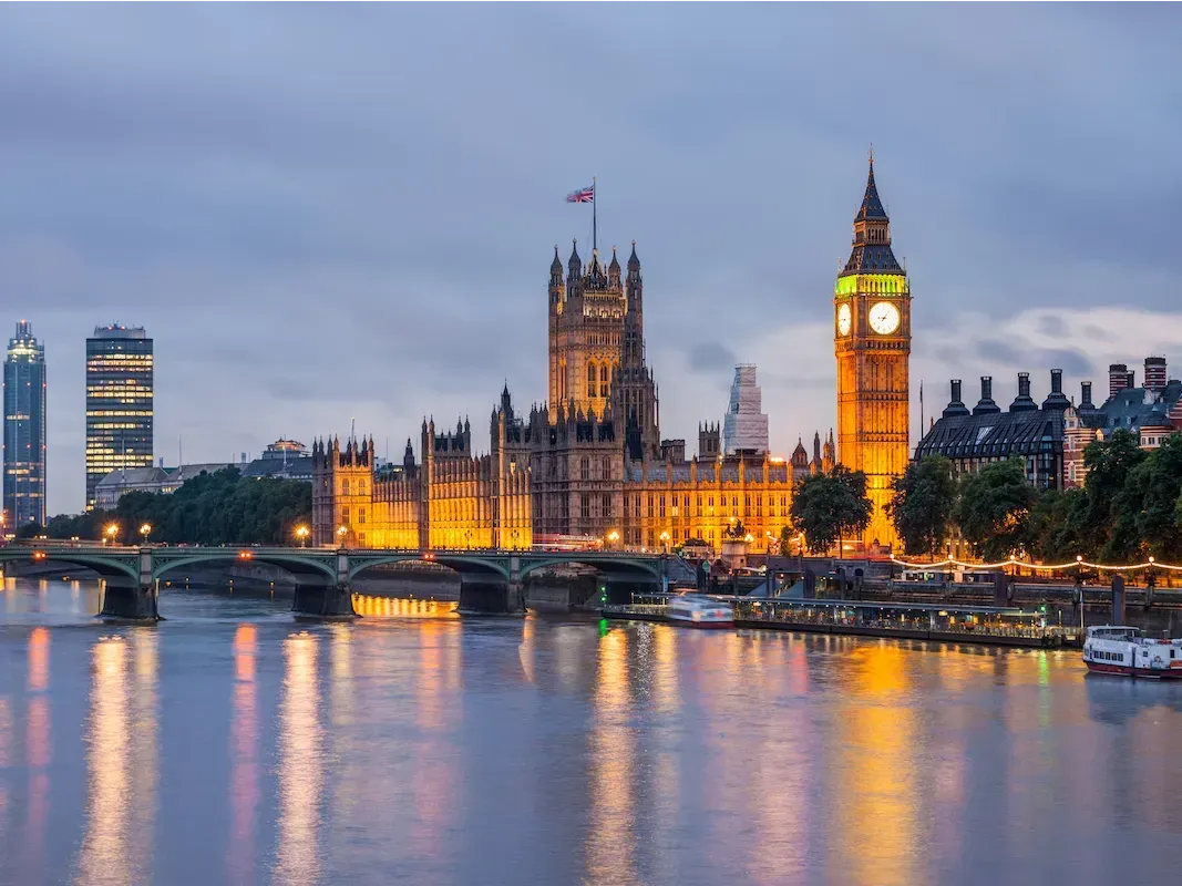 View of the Houses of Parliament and Big Ben illuminated at dusk along the River Thames in London.