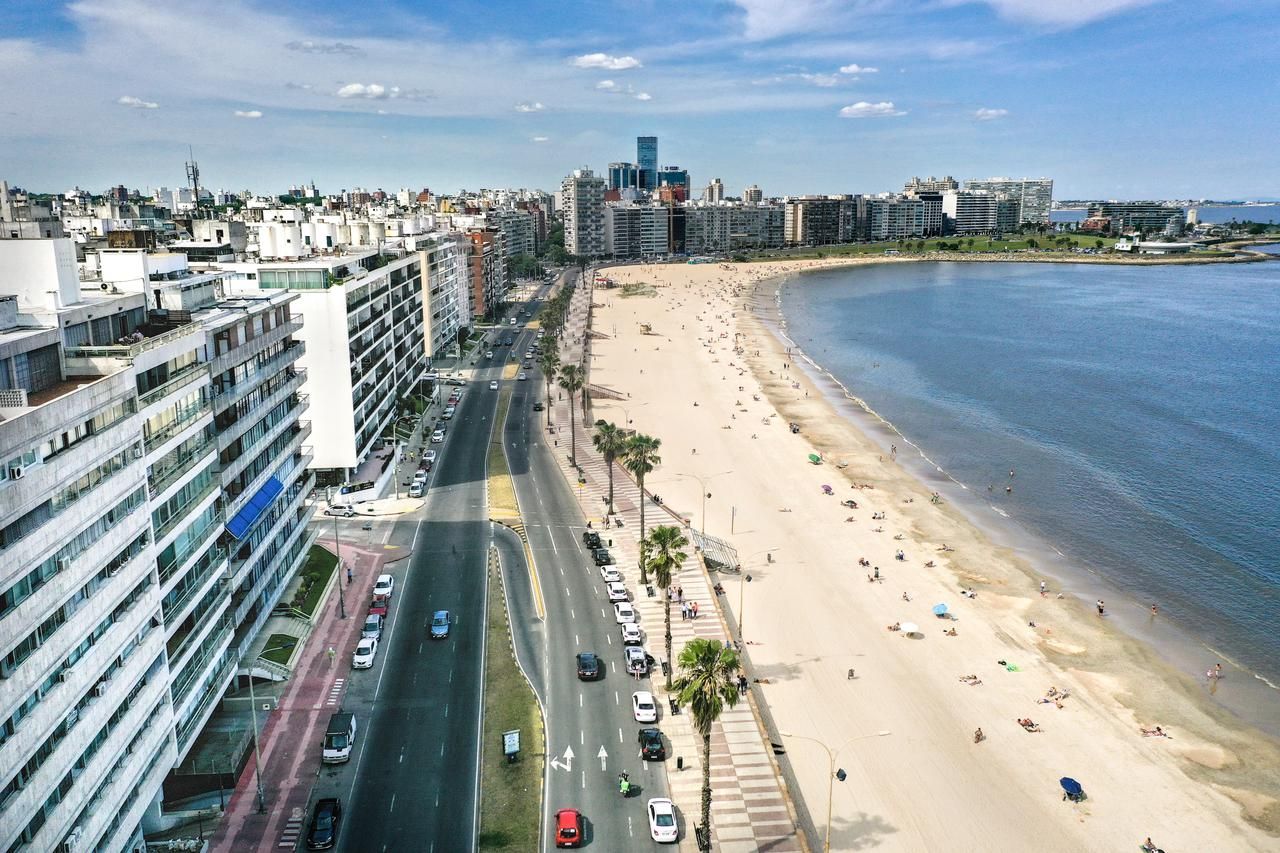 Aerial view of a beach in Montevideo, Uruguay. A wide beach with people, a coastal road with cars, and city buildings.