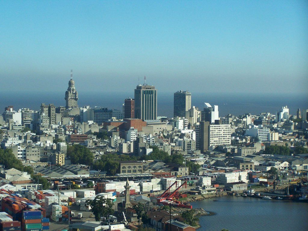 Panoramic view of Montevideo, Uruguay, with a harbor in the foreground and a cityscape of diverse buildings under a blue sky.