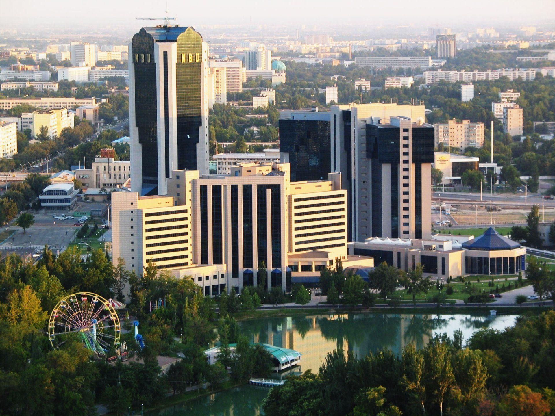 Cityscape with high-rise buildings, including a tall skyscraper, reflected in a lake. Park with Ferris wheel in foreground.
