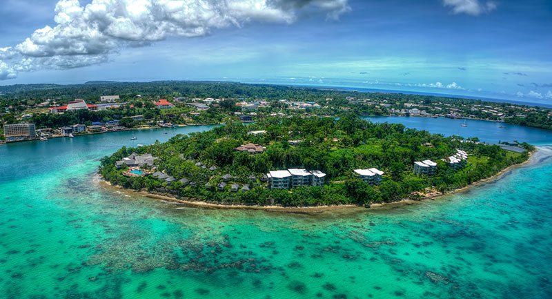 Aerial view of an island surrounded by turquoise water and lush greenery, with buildings and a city visible in the background.