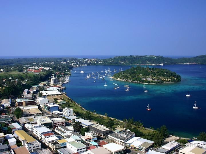 Aerial view of Port Vila, Vanuatu, with a town, harbor, and island under a clear blue sky. Sailboats dot the turquoise water.