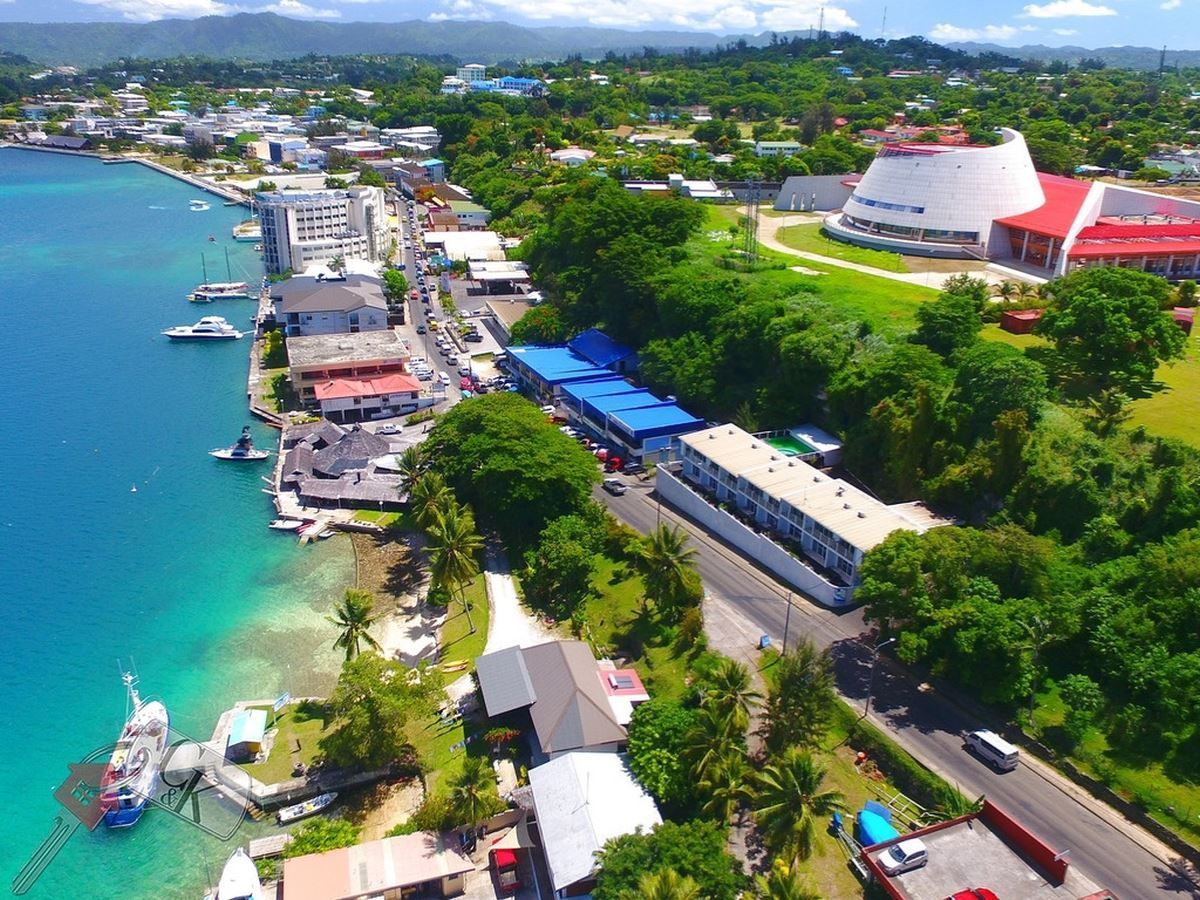 Aerial view of a coastal town with turquoise water. Buildings line the shore; a large, white, domed structure is on a grassy hill.
