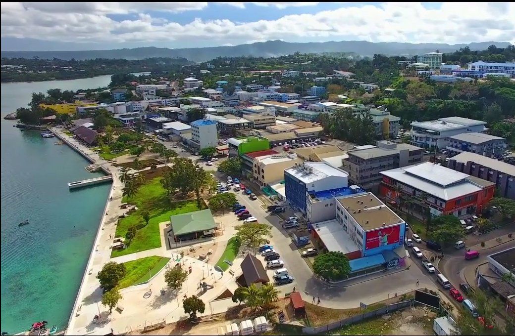 Aerial view of a coastal town with buildings, roads, and a waterfront park. Clear blue water and lush green hills are in the background.